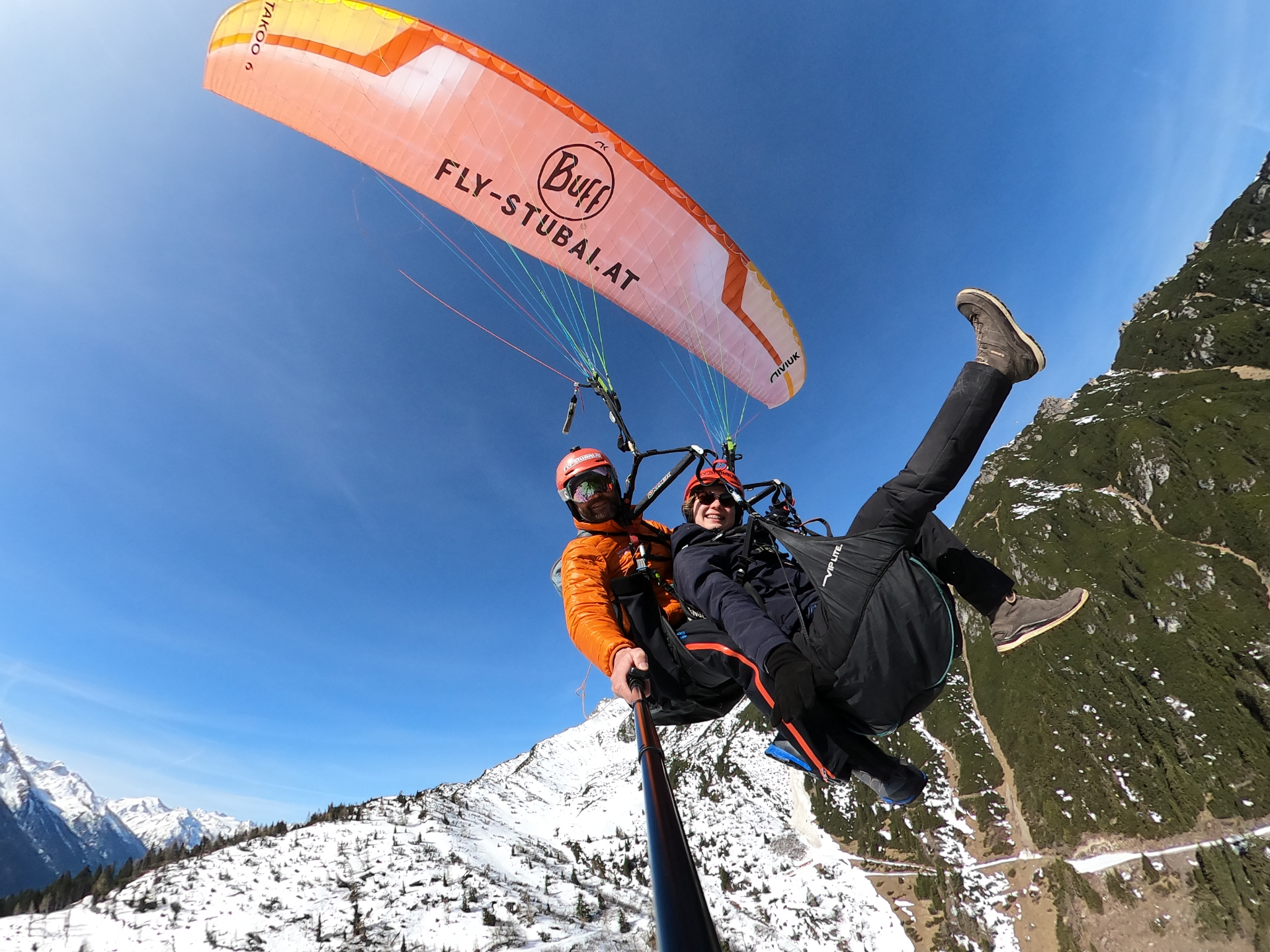 Winterlicher Tandemflug mit schneebedeckten Bergen und orangefarbenem Gleitschirm in Tirol
